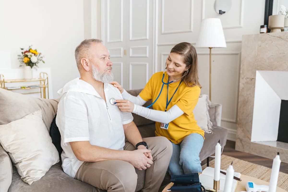 A nurse checking an older man with a stethoscope in a living room.